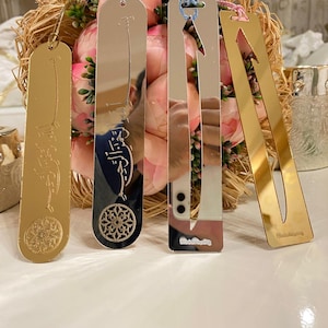 May include: Four silver and gold bookmarks with decorative designs and tassels. The bookmarks are on a white surface with a bouquet of pink flowers in the background.