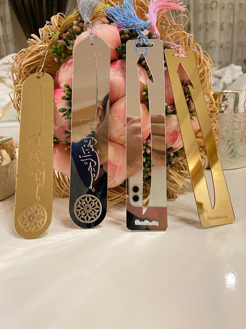 May include: Four silver and gold bookmarks with decorative designs and tassels. The bookmarks are on a white surface with a bouquet of pink flowers in the background.