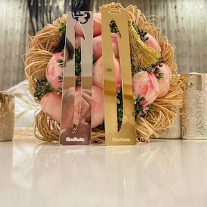May include: Two silver and gold bookmarks with tassels. The bookmarks are shaped like arrows and have a decorative design. The bookmarks are on a table with a bouquet of pink flowers.