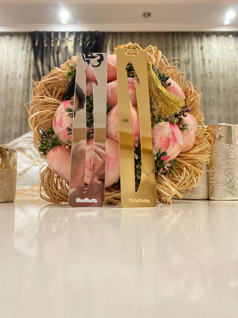 May include: Two silver and gold bookmarks with tassels. The bookmarks are shaped like arrows and have a decorative design. The bookmarks are on a table with a bouquet of pink flowers.