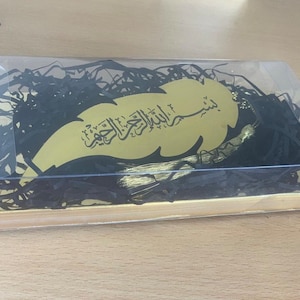 May include: A gold leaf-shaped bookmark with black Arabic calligraphy and a gold tassel. The bookmark is packaged in a clear plastic box with black shredded paper.
