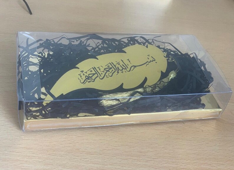 May include: A gold leaf-shaped bookmark with black Arabic calligraphy and a gold tassel. The bookmark is packaged in a clear plastic box with black shredded paper.