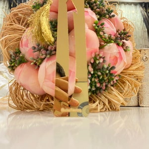 May include: A gold bookmark with a tassel, featuring a pointed arrow design. The bookmark is resting on a bed of straw and pink flowers.