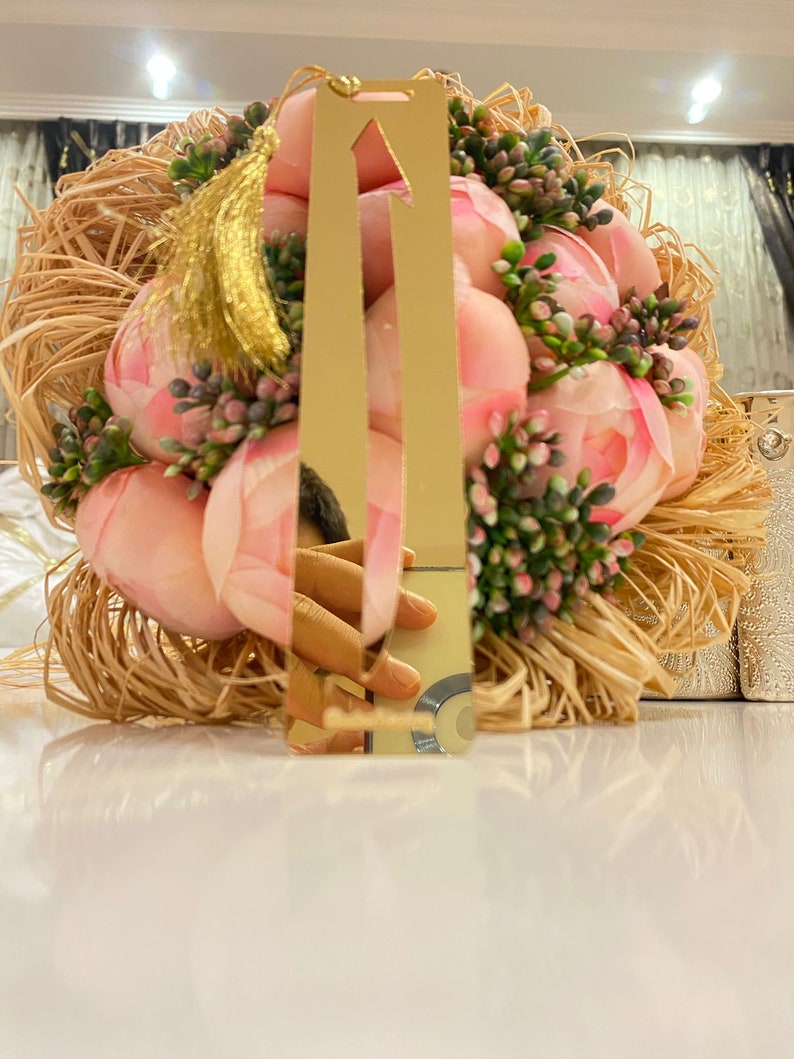 May include: A gold bookmark with a tassel, featuring a pointed arrow design. The bookmark is resting on a bed of straw and pink flowers.
