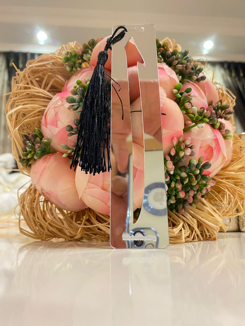 May include: A silver bookmark with a black tassel. The bookmark is shaped like an arrow and has a reflective surface.