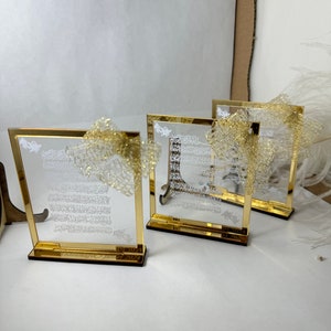 May include: Three gold-framed glass plaques with white Arabic calligraphy and a gold glitter overlay. The plaques have a wooden base and are arranged in a row on a white surface.