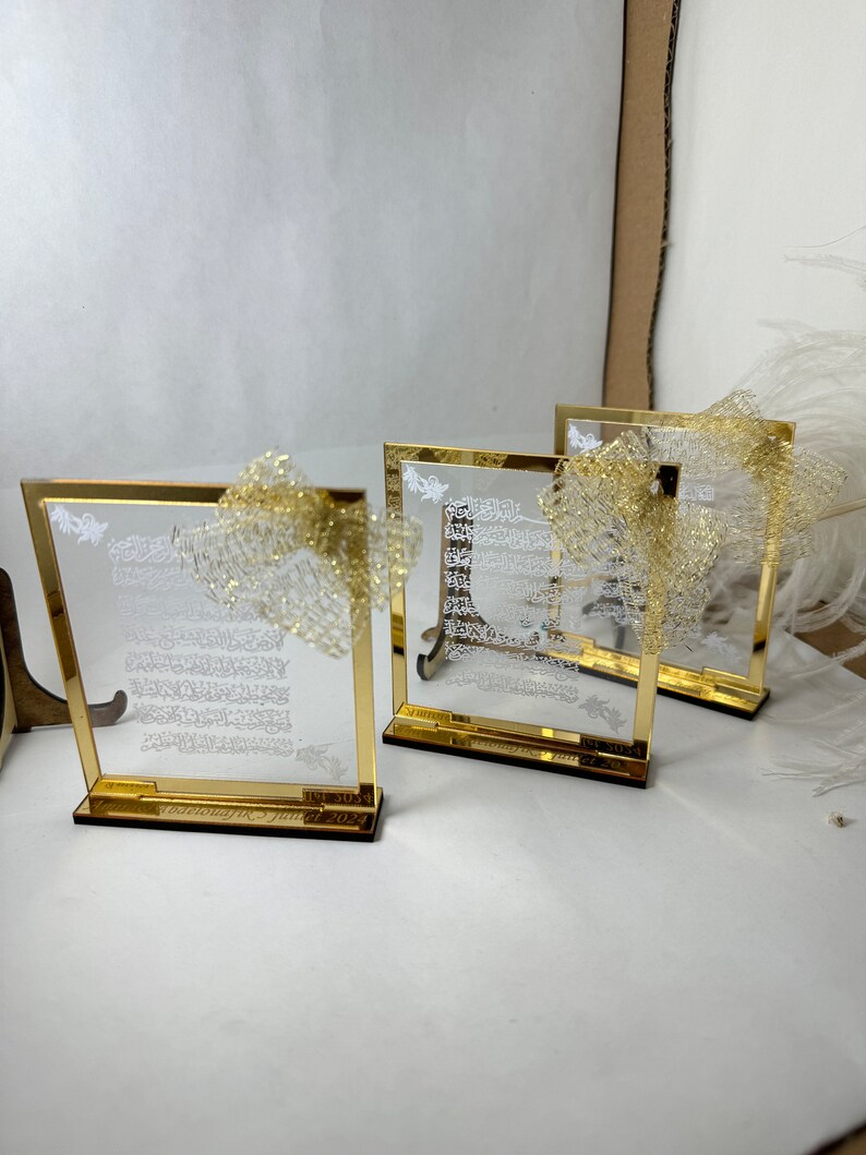 May include: Three gold-framed glass plaques with white Arabic calligraphy and a gold glitter overlay. The plaques have a wooden base and are arranged in a row on a white surface.