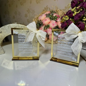 May include: Two gold-framed glass plaques with white Arabic calligraphy inside. Each plaque is on a gold stand and has a white satin bow on top.