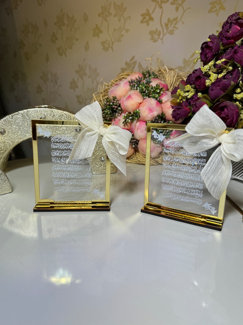 May include: Two gold-framed glass plaques with white Arabic calligraphy inside. Each plaque is on a gold stand and has a white satin bow on top.