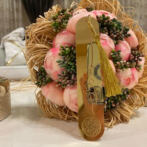 May include: A gold bookmark with a tassel and a decorative design. The bookmark is resting on a bouquet of pink peonies.