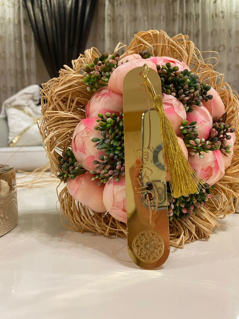 May include: A gold bookmark with a tassel and a decorative design. The bookmark is resting on a bouquet of pink peonies.