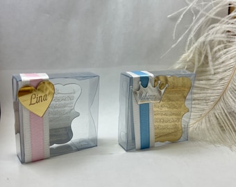 May include: Two clear plastic boxes with gold-colored metal ornaments inside. The box on the left has a pink ribbon and a heart-shaped ornament with the name "Lina" on it. The box on the right has a blue ribbon and a crown-shaped ornament with the name "Mohammed" on it.