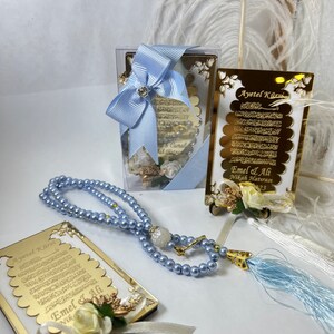 May include: A set of wedding favours featuring a gold-framed, white card with Arabic calligraphy and a blue tassel. The card is decorated with a white flower and a white ribbon. The set also includes a blue prayer bead necklace and a small, clear box with a blue ribbon.