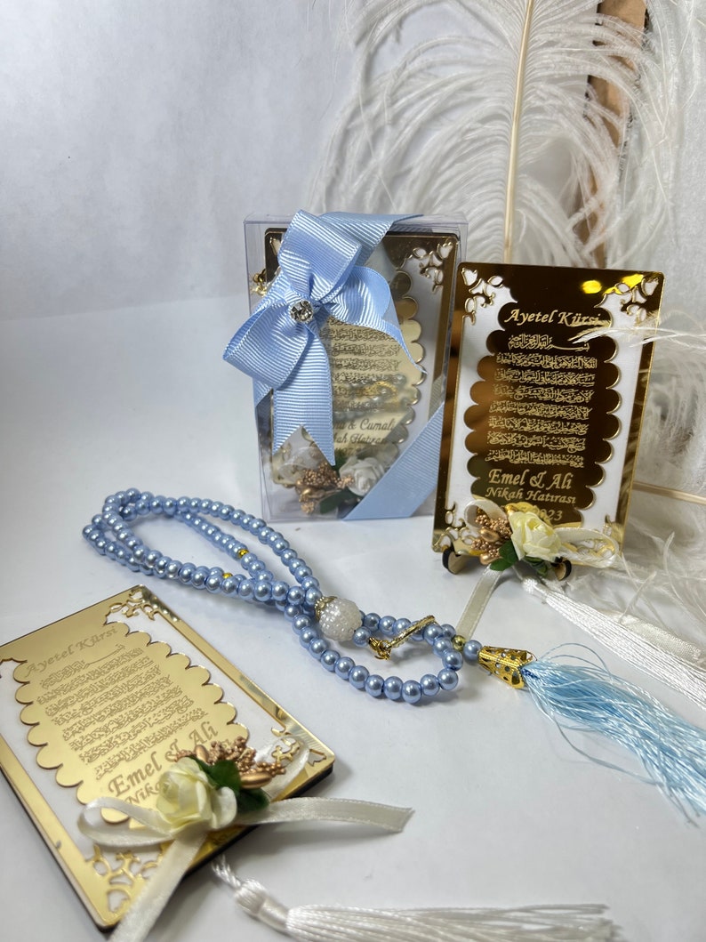 May include: A set of wedding favours featuring a gold-framed, white card with Arabic calligraphy and a blue tassel. The card is decorated with a white flower and a white ribbon. The set also includes a blue prayer bead necklace and a small, clear box with a blue ribbon.