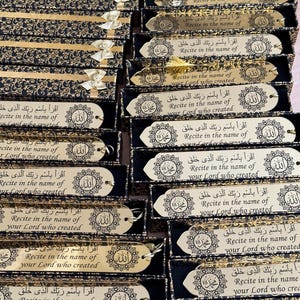 May include: A close-up of several gold bookmarks with an Arabic inscription and a decorative design. The bookmarks are packaged in black boxes with gold accents.