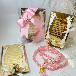 May include: A pink and gold Islamic prayer set with a small framed verse, a larger framed verse, and a pink prayer bead necklace with a tassel.