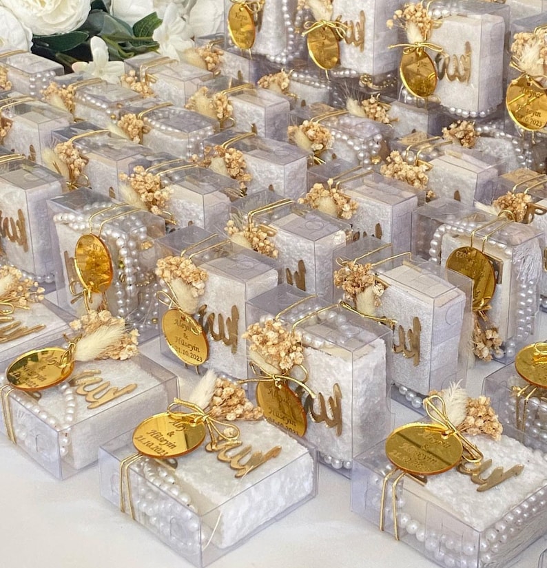 May include: A collection of small, clear boxes filled with white velvet and decorated with gold accents. Each box features a small, white, wooden plaque with the word "Allah" written in Arabic calligraphy. The boxes are adorned with white pearl beads and dried flowers.