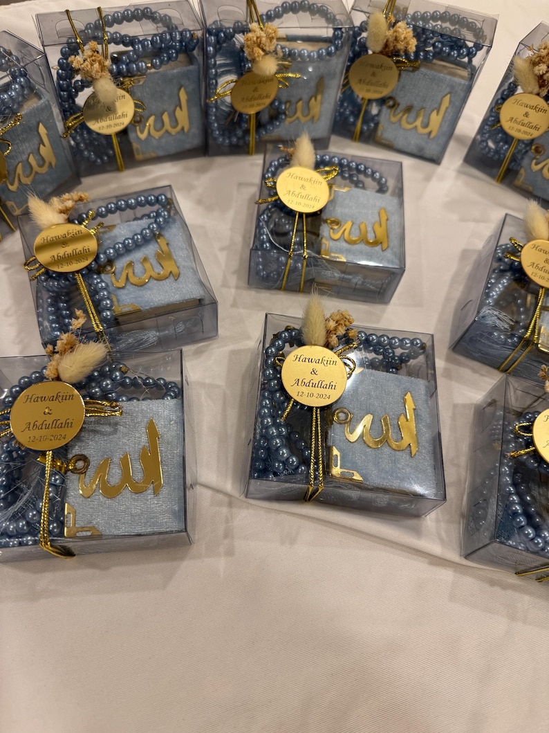 May include: A collection of small, clear boxes containing blue prayer beads and a small blue fabric book with gold lettering. The boxes are decorated with a small white flower and a gold tag with the text "Hawakin & Abdullahi 12-10-2024".
