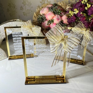 May include: Three gold-framed glass plaques with Arabic calligraphy and a gold ribbon bow. The plaques are on a white surface with a bouquet of pink flowers and a purple flower arrangement in the background. The bottom plaque has the inscription "Seyma Yra Demirci 12.01.2024".