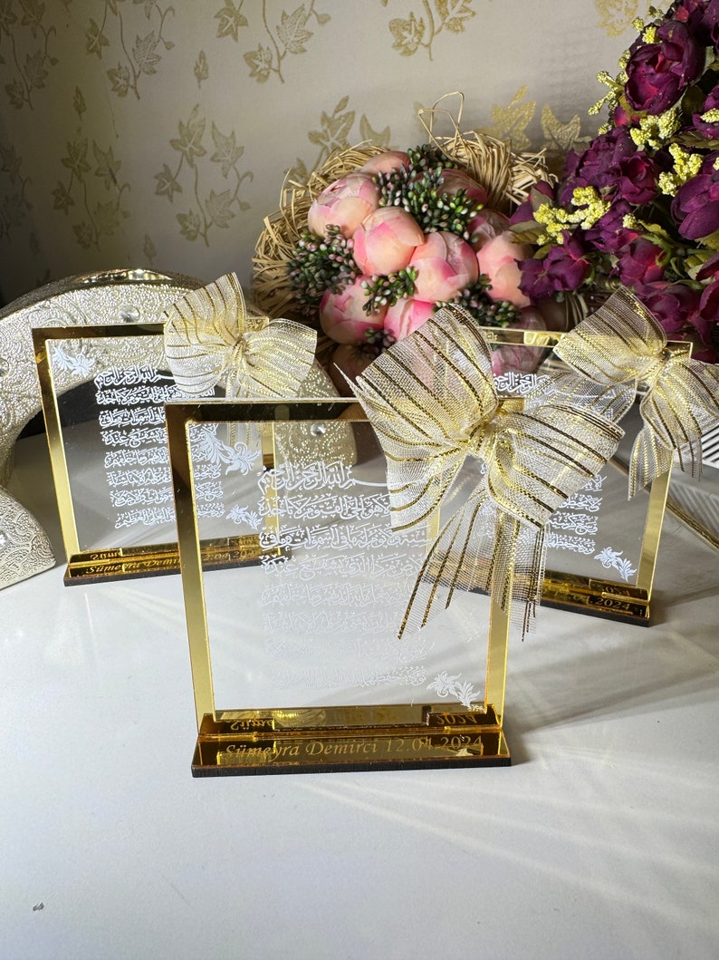 May include: Three gold-framed glass plaques with Arabic calligraphy and a gold ribbon bow. The plaques are on a white surface with a bouquet of pink flowers and a purple flower arrangement in the background. The bottom plaque has the inscription "Seyma Yra Demirci 12.01.2024".