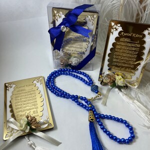 May include: A set of three gold-framed Islamic prayer cards with blue and white tassels. The cards feature the Arabic text of the Ayatul Kursi, a verse from the Quran. The cards are packaged in a clear plastic box with a blue ribbon. The set also includes a blue prayer bead necklace with a tassel.