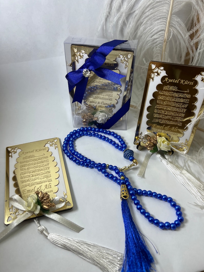 May include: A set of three gold-framed Islamic prayer cards with blue and white tassels. The cards feature the Arabic text of the Ayatul Kursi, a verse from the Quran. The cards are packaged in a clear plastic box with a blue ribbon. The set also includes a blue prayer bead necklace with a tassel.