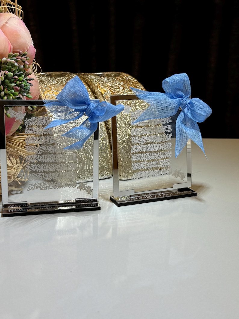 May include: Two clear glass frames with engraved text and blue bows. The frames are on a white surface. The text on the frames reads "Welcome Baby A" and "Welcome Baby Z".