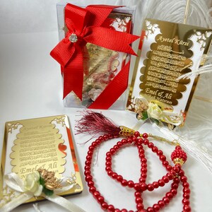 May include: Two gold-framed plaques with Arabic calligraphy and the text "Ayet el Kirsi" and "Emel & Ali" with a white flower and a red ribbon. A red prayer bead necklace with gold accents.