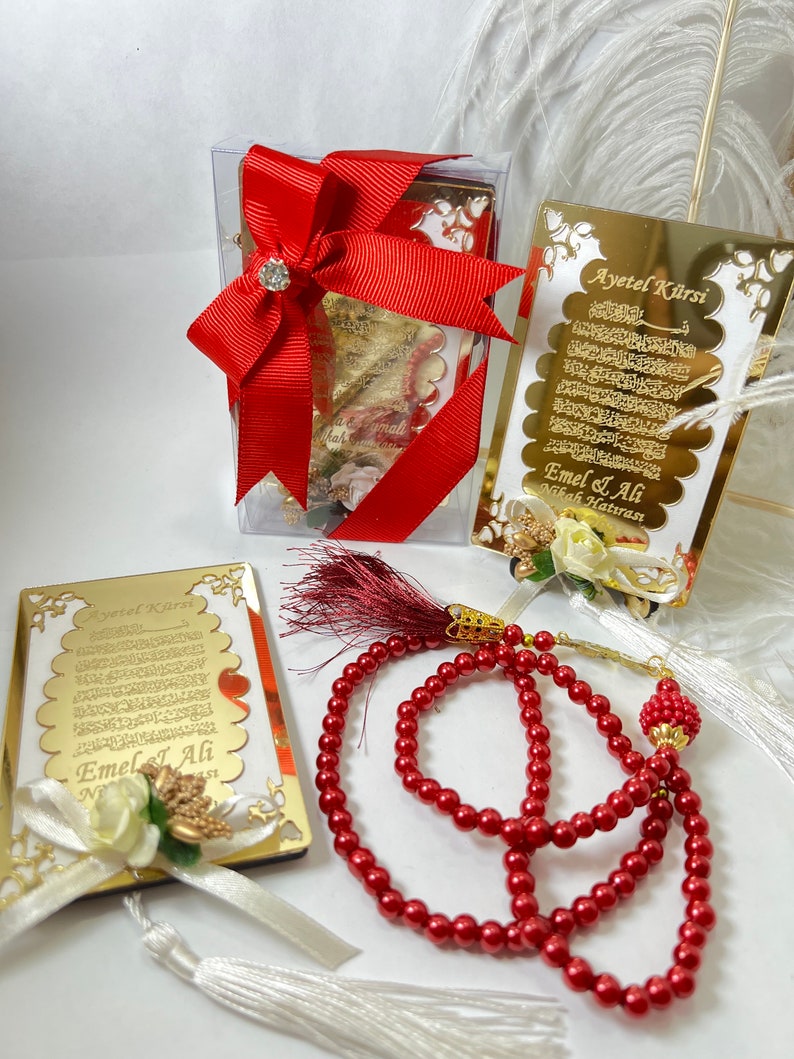 May include: Two gold-framed plaques with Arabic calligraphy and the text "Ayet el Kirsi" and "Emel & Ali" with a white flower and a red ribbon. A red prayer bead necklace with gold accents.