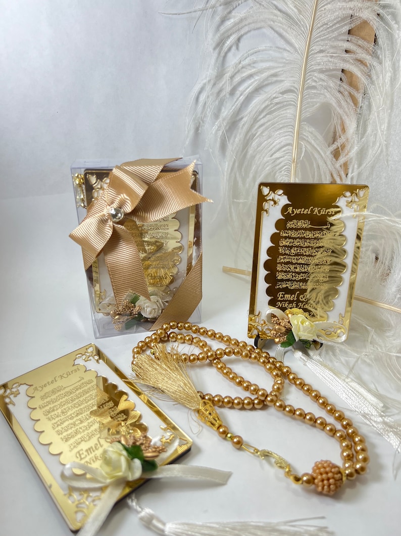 May include: A gold-coloured framed print of the Ayatul Kursi, a verse from the Quran, with a white background and gold lettering. The print is in a clear plastic box with a gold ribbon. A gold-coloured prayer bead necklace with a gold tassel is also included. The print is decorated with white flowers and a gold ribbon.