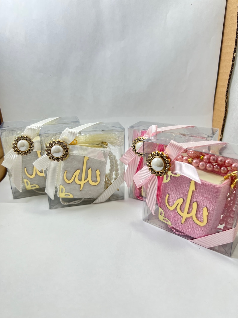 May include: Three clear gift boxes with pink and white velvet books inside. Each box has a gold ribbon with a pearl and gold accent. The books have the word "Allah" written in gold on the front.