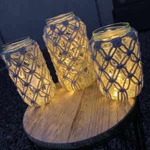 Könnte beinhalten: Drei Glasgläser mit weißen Makramee-Bezügen sind von innen beleuchtet und erzeugen ein warmes Leuchten. Die Gläser stehen auf einem Holztisch mit einem schwarzen Metallfuß.