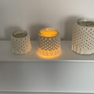Könnte beinhalten: Drei Kerzenhalter aus Glas mit cremefarbenen Makramee-Hüllen. Der mittlere Kerzenhalter ist beleuchtet und strahlt ein warmes Licht aus. Die Makramee-Hüllen haben unten eine Fransen. Die Kerzenhalter stehen auf einem weißen Regal.
