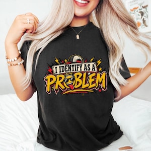May include: A black short-sleeved t-shirt featuring a graphic design with the text I IDENTIFY AS A PROBLEM in bold yellow letters. The design includes a yellow emoji wearing sunglasses and a baseball cap, surrounded by red and yellow lightning bolts.