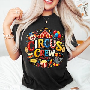 Op de afbeelding: Zwart t-shirt met een kleurrijk circusthema. Het ontwerp bevat een clown, circustent, ballonnen en de woorden "CIRCUS CREW" in een speels lettertype. Extra elementen zijn een trommel, popcorn en een zeehond.