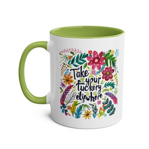 Puede incluir: Taza de cerámica blanca con borde verde. La taza presenta un diseño floral colorido y el texto "Take your f*ckery elsewhere".