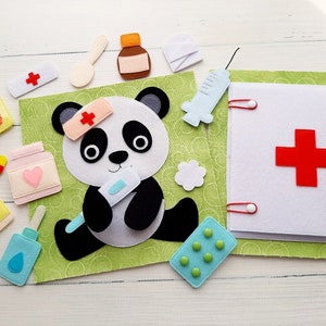 Puede incluir: Un libro de fieltro para niños con un oso panda que lleva un gorro de médico y sostiene un termómetro. El libro tiene páginas con varios suministros médicos, incluyendo píldoras, vendas y una jeringa. El libro está sobre una superficie de madera blanca.