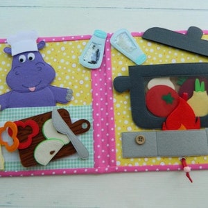 Könnte beinhalten: Ein Filzbuch für Kinder mit einem lila Hippopotamus auf der linken Seite und einem schwarzen Topf mit einer roten Tomate, einer Zwiebel und anderem Gemüse auf der rechten Seite. Der Topf hat einen Deckel, der angehoben werden kann.