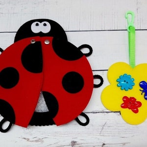 Peut inclure: Une coccinelle rouge avec des points noirs et une fleur jaune avec des boutons colorés. La coccinelle est en feutre et a une fermeture velcro. La fleur est également en feutre et a une boucle pour l'accrocher.