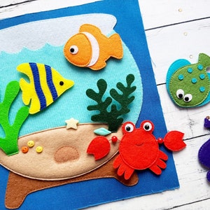 Puede incluir: Conjunto de juguetes de fieltro que incluye una pecera azul con un pez amarillo y azul, algas verdes, un cangrejo rojo, un pez globo verde y un caballito de mar morado. La pecera tiene una base marrón y un borde blanco.