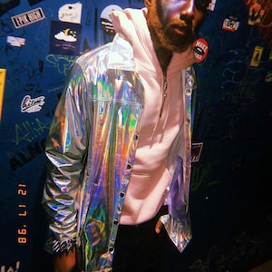 May include: A silver iridescent jacket with a button-up front. The jacket has a shiny, reflective surface that changes colours. The jacket is worn over a light pink hooded sweatshirt. The background is a dark blue wall with graffiti and stickers.