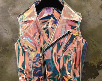 Puede incluir: Chaqueta iridiscente sin mangas con cierre de cremallera. La chaqueta tiene cuello y un acabado holográfico brillante, que refleja múltiples colores. La prenda está colgada de una percha negra sobre un fondo gris.
