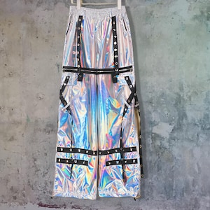 May include: Holographic silver trousers with black straps and grommet details. The trousers have a wide-leg design and a zip closure. The material reflects a rainbow of colours, creating a futuristic look. The trousers are hanging against a grey concrete wall.