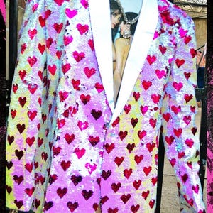 Puede incluir: Un blazer de lentejuelas con solapas blancas, cubierto de lentejuelas rojas y rosas en forma de corazón. Se lleva sobre una camiseta azul claro con un estampado gráfico. El conjunto es audaz y llamativo.