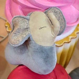 May include: A gray plush elephant toy with a beige face and closed eyes, sitting on top of a pink and white cupcake.