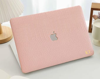 May include: A light pink laptop hard shell case with a textured crocodile skin pattern. The case features a centered cutout for the Apple logo and the gold-colored initials KT. printed in the bottom right corner of the cover.