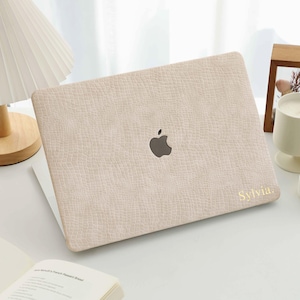 Può includere: Una custodia per laptop beige chiaro con un motivo strutturato e il logo Apple. Il nome "Sylvia" è stampato in oro in basso a destra. Il laptop è aperto, rivelando uno schermo bianco.