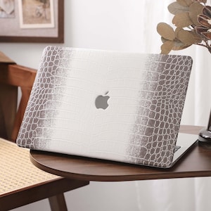 May include: A laptop with a white and brown crocodile-textured case. The case has a gradient design, transitioning from white to brown. The Apple logo is visible on the laptop's lid. The laptop is on a dark wooden table.