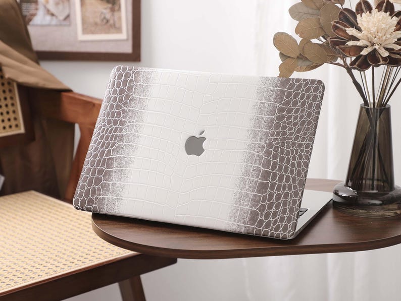 May include: A laptop with a white and brown crocodile-textured case. The case has a gradient design, transitioning from white to brown. The Apple logo is visible on the laptop's lid. The laptop is on a dark wooden table.