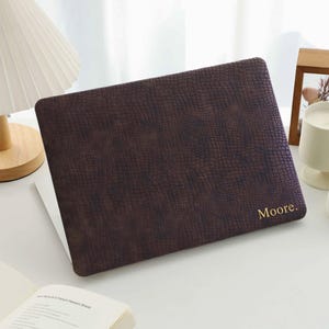 May include: A dark brown laptop cover with a textured crocodile skin pattern and the name Moore. printed in gold lettering on the bottom right corner. The cover is rectangular with rounded corners and fits securely over the laptop lid.